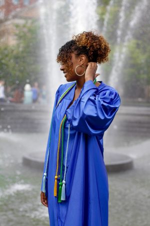 A woman in a blue graduation gown stands near a fountain, adjusting her hair.