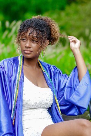 Woman in a blue graduation gown adjusting her hair outdoors.