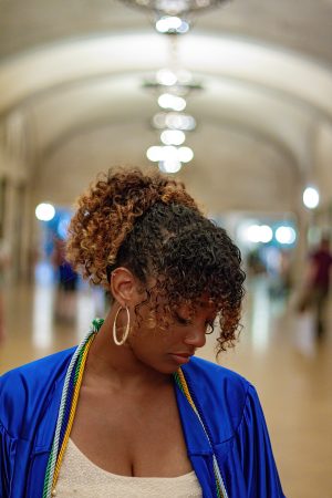 Young woman with curly hair in a blue jacket, looking down in a softly lit arched hallway.