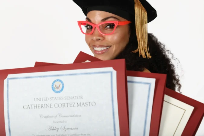 Graduate smiling with diploma and red glasses in black cap and gown.