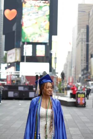 A smiling graduate in a blue cap and gown in Times Square.