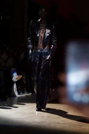 Model in a shiny black suit walking on a runway during a fashion show.