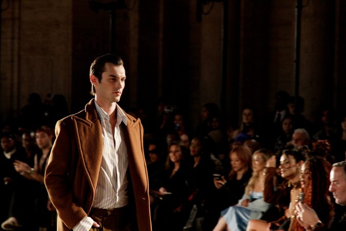Male model wearing a brown jacket on runway during fashion show.