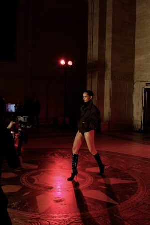 Model walking on a runway in a dimly lit venue wearing a dark outfit and boots.