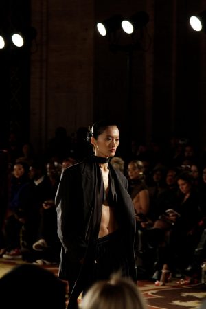 Model walks runway wearing a dark, draped outfit with a serious expression.