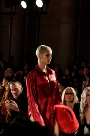 Model in a red outfit walks runway with attentive audience.