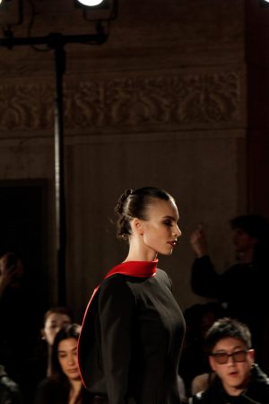 Profile view of a model in a black and red outfit on a dimly lit runway.