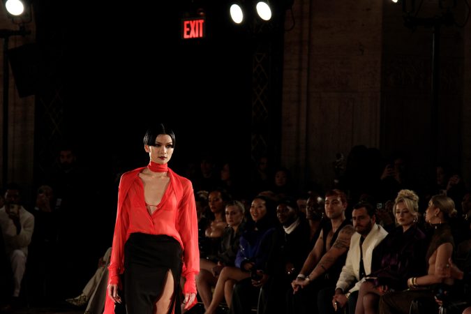 Model walking on runway in red top and black skirt at fashion show.