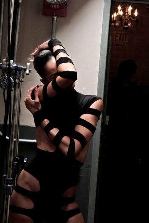 Person wrapped in black bands posing in low light.